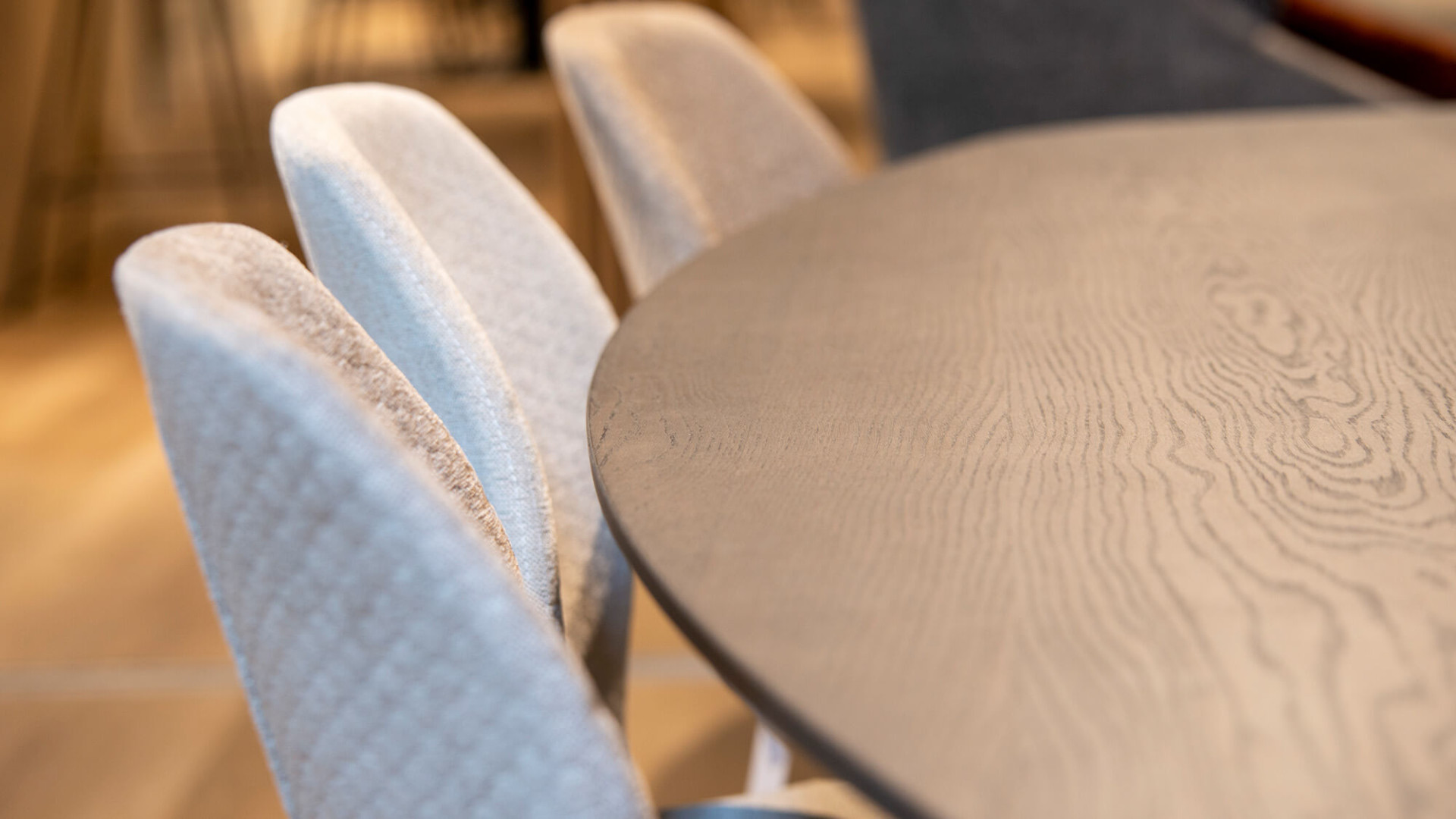 Close-up van een moderne tafel met een gladde, donkere houten bovenkant, omringd door comfortabele, lichtgrijze gestoffeerde stoelen, met een warme vloer op de achtergrond.